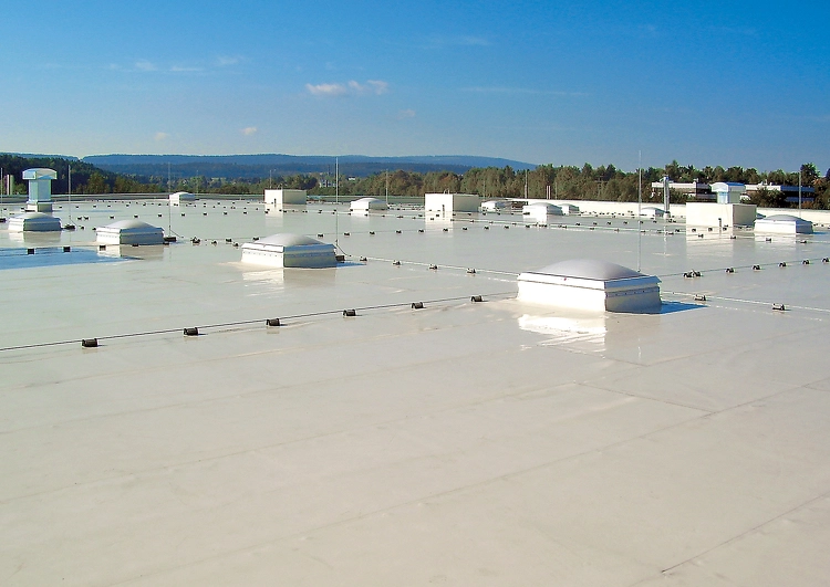 grande surface de toiture plate couverte en membrane TPO, avec de nombreux puits de lumière et sortie de ventilation, végétation et reliefs à l'arrière plan.