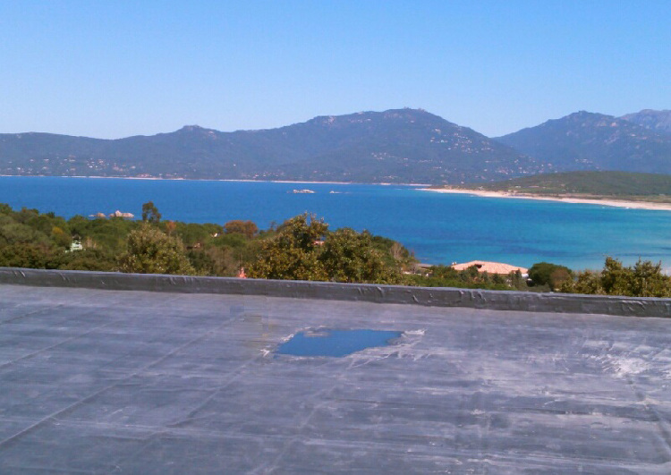 au premier plan une toiture plate avec acrotère, couverture en membrane EPDM, à l'arrière plan vue sur la mer, la cote avec une plage à droite et des montagnes au fond.