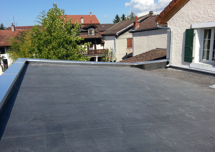 toiture en pente douce couverte en membrane EPDM, bordée à droite et au fond par des acrotères avec couvertines, à droite un mur avec une fenêtre aux volets verts, à l'arrière plan d'autres maisons et un arbre, en zone urbaine.